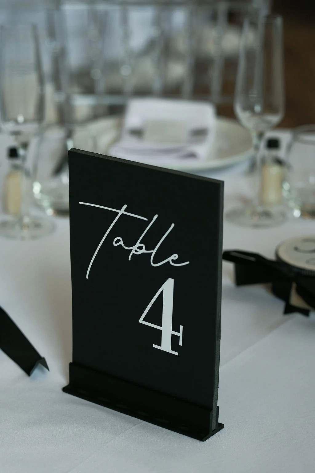 A sleek, modern black table number sign with white calligraphy sits on a crisp white table linen during a wedding reception.