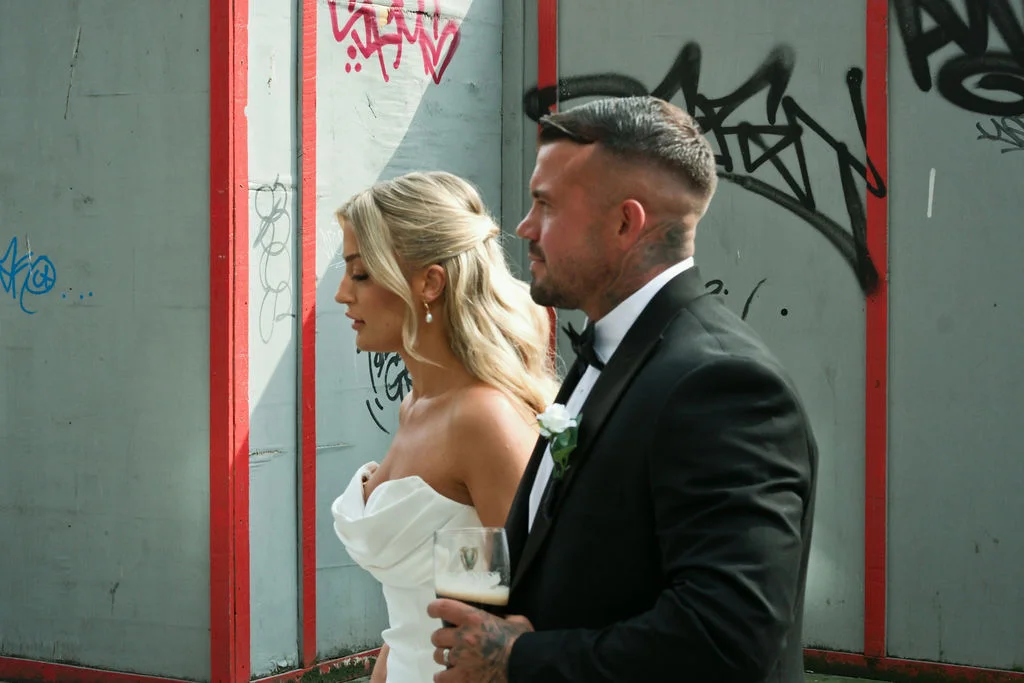 A bride and groom walk together against an urban graffiti backdrop, with the groom wearing a classic black tuxedo and holding a pint of beer.