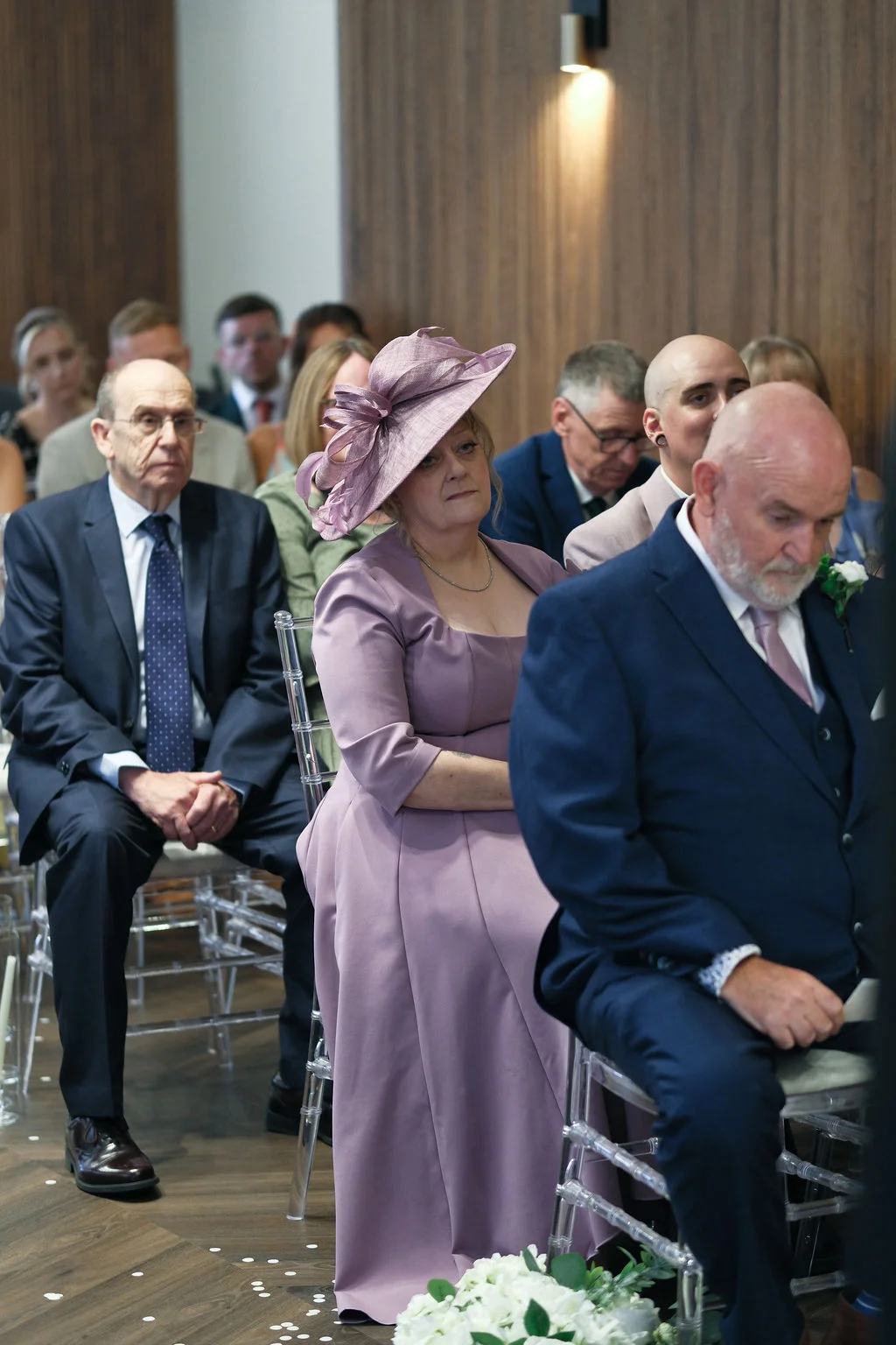 Wedding guests seated on clear acrylic chairs during a ceremony, with a woman wearing a lavender dress and matching fascinator prominently featured in the middle row.