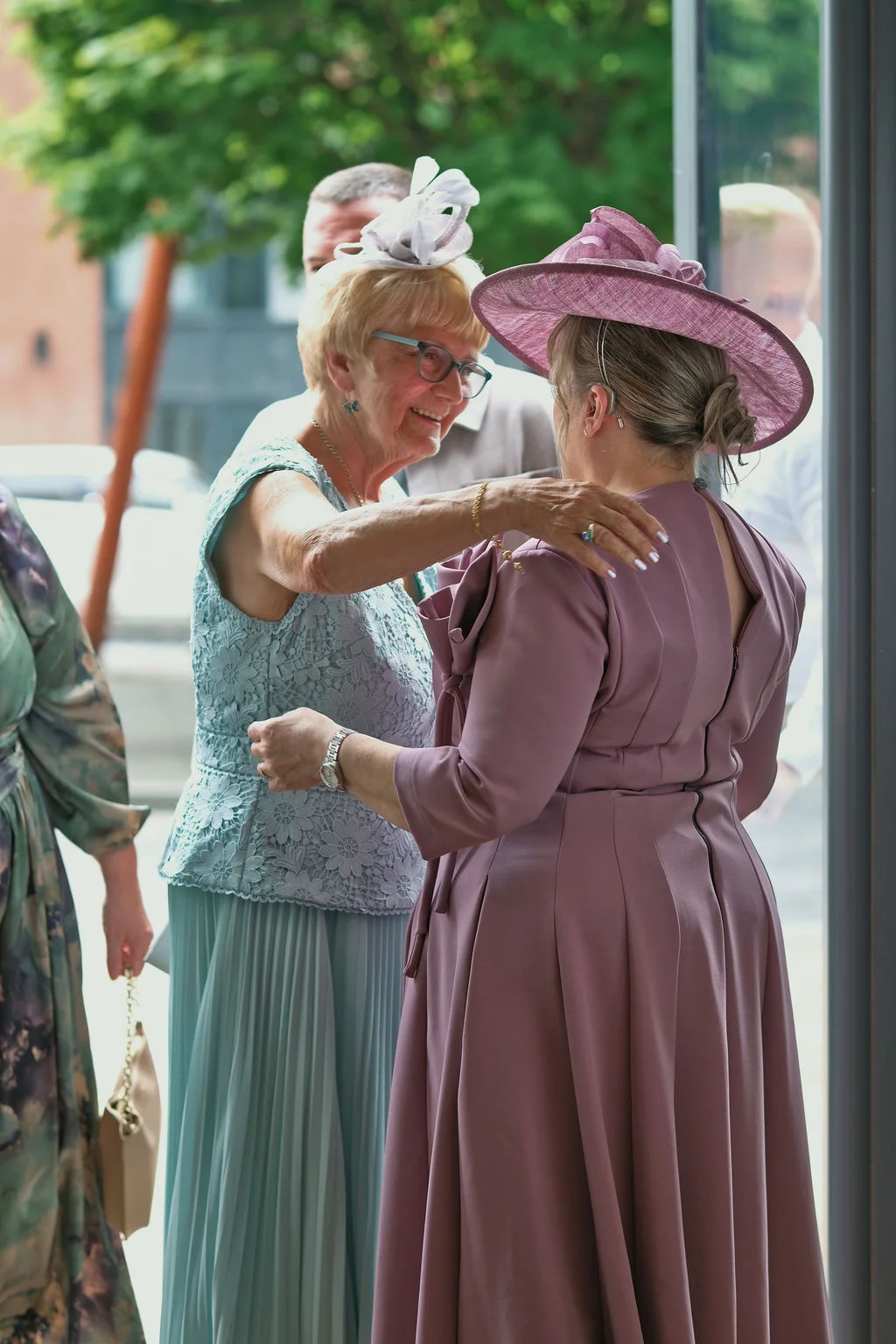 Two older female wedding guests in elegant dresses and matching fascinators warmly embrace each other near an entrance.