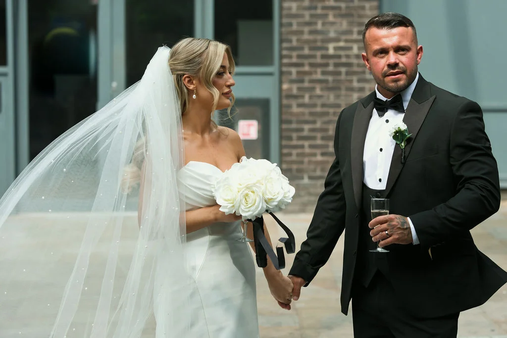 A newlywed couple walking hand in hand outdoors, with the bride wearing a strapless white gown and pearl veil, and the groom in a classic black tuxedo holding a glass of champagne.