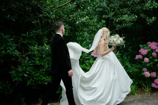 A newlywed couple walks through a lush woodland setting, with the bride wearing a flowing satin gown and a dramatic veil catching the breeze.