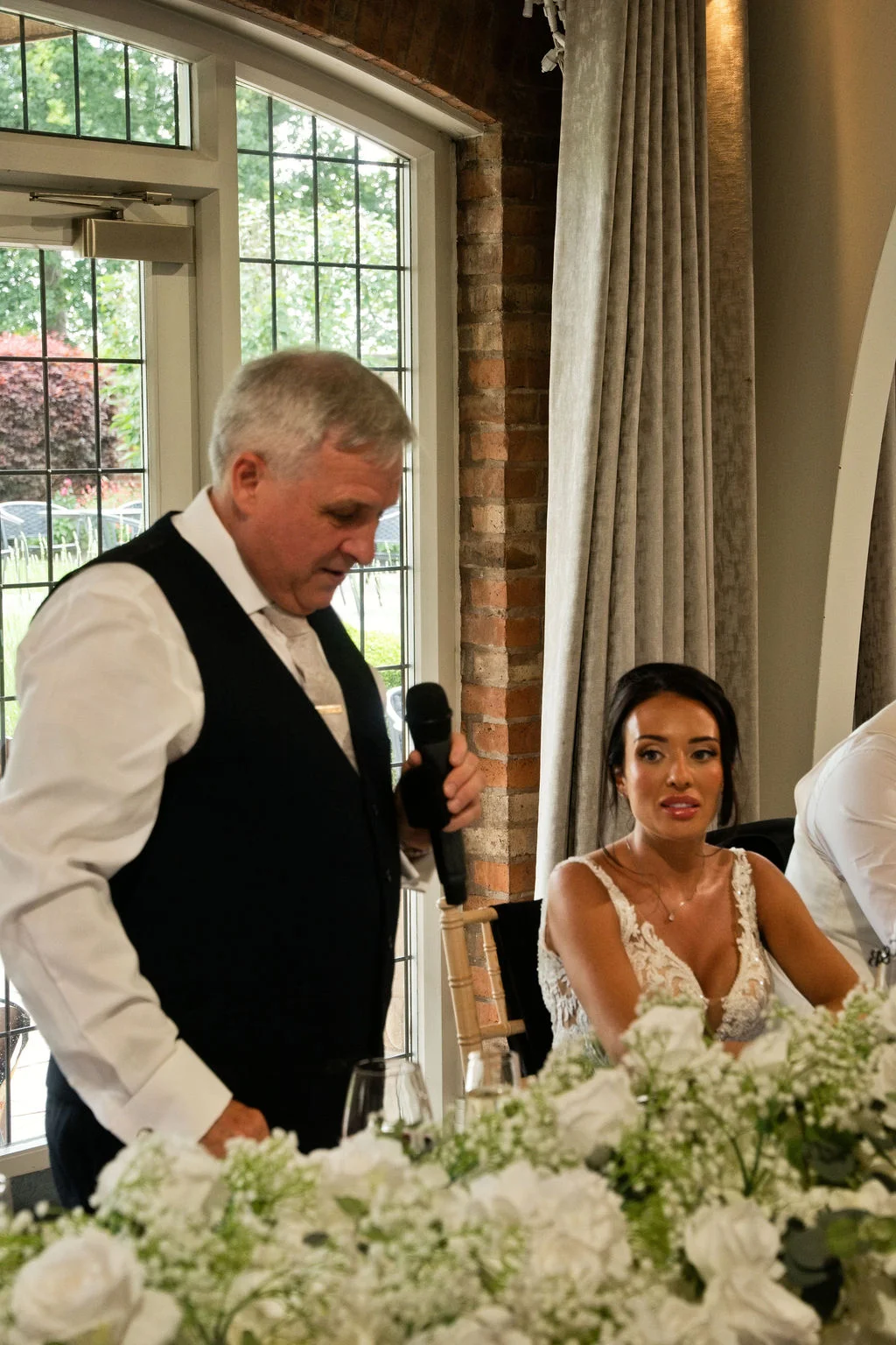 A father of the bride delivers a speech holding a microphone at the top table while the bride listens attentively beside a white floral centrepiece.
