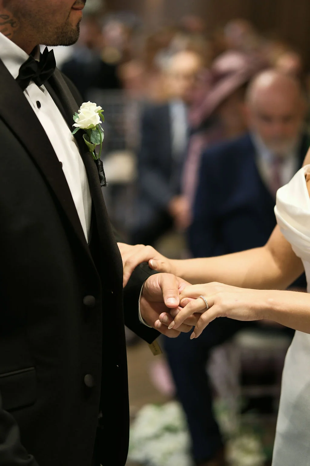 The bride and groom hold hands during their wedding ceremony, showing a sparkling engagement ring and a black tuxedo with a white rose boutonniere.