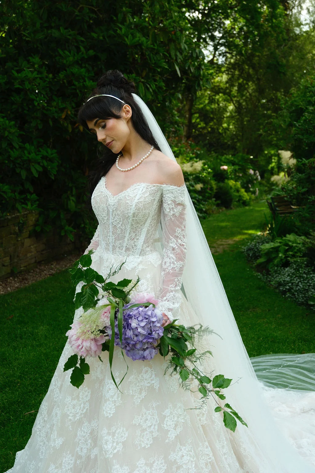 A stunning bride with dark hair wearing an off-the-shoulder lace gown and pearl necklace stands in a lush garden holding a cascading bouquet of pink and purple flowers.