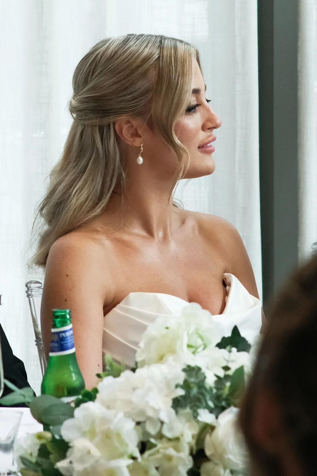 A profile shot of a blonde bride with a half-up hairstyle and pearl drop earrings, seated at a reception table adorned with white floral centerpieces.