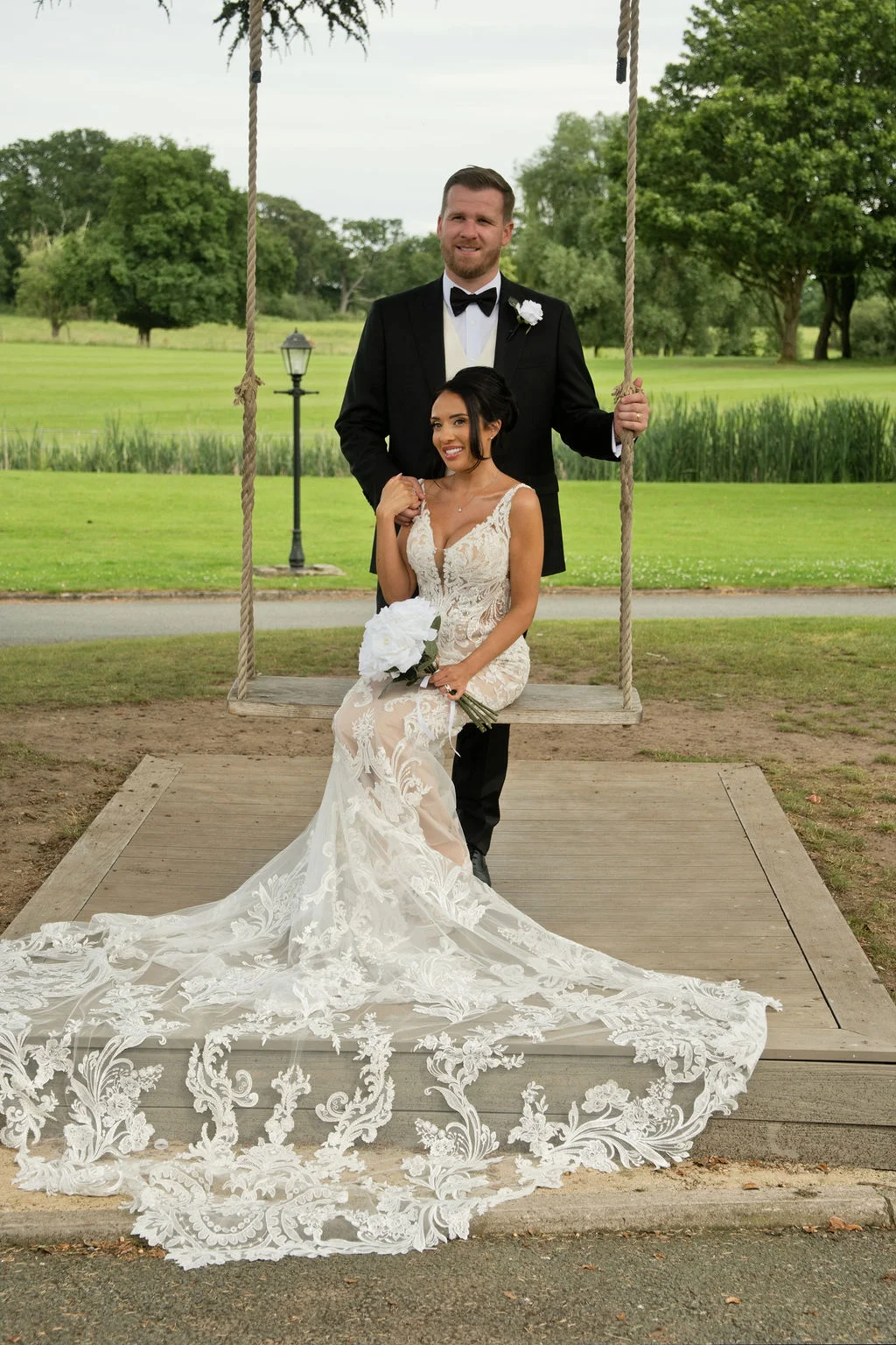 A newlywed couple poses outdoors with the bride seated on a rustic rope swing wearing a lace mermaid gown with a long train, while the groom stands behind her in a black tuxedo.