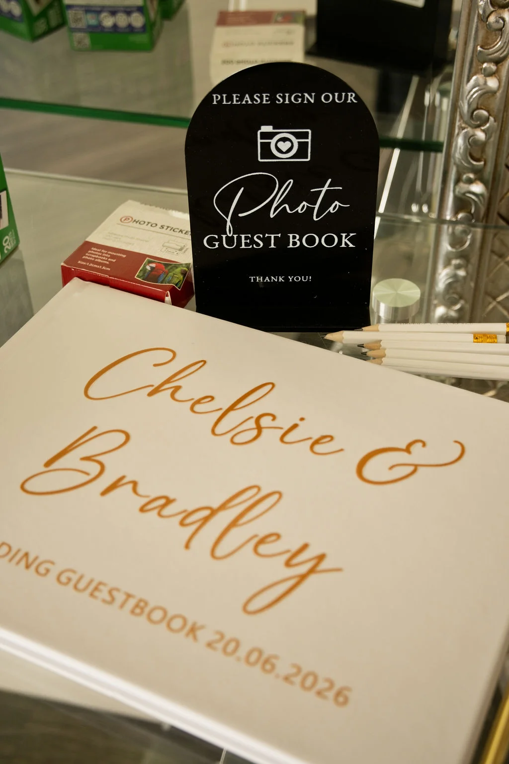 A personalised wedding photo guest book with gold lettering lies on a glass table alongside a matching black acrylic sign and white pencils.