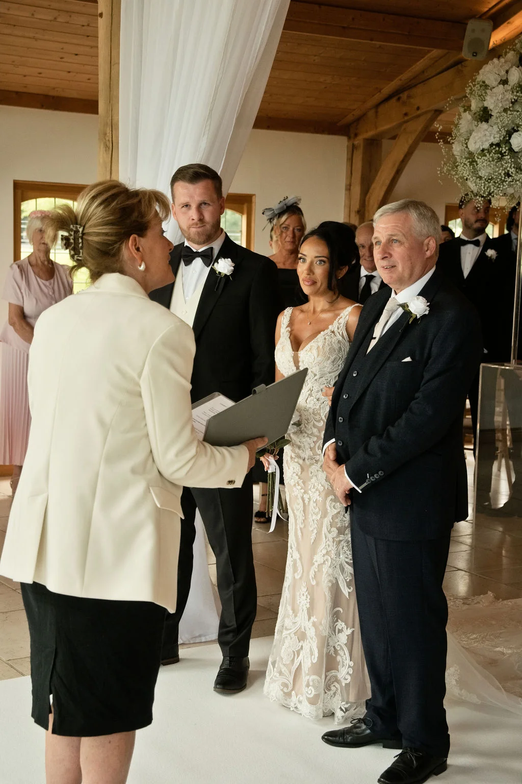 The bride and groom stand together during a civil ceremony inside a light-filled barn venue with wooden rafters, surrounded by family and guests.