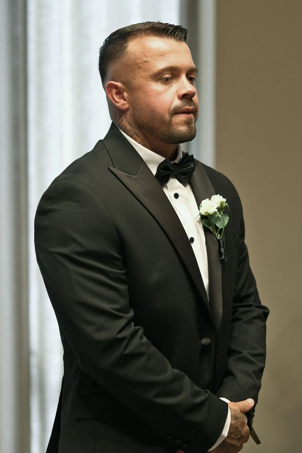 A sharp groom stands waiting at the altar wearing a classic black tuxedo, bow tie, and a white rose buttonhole.