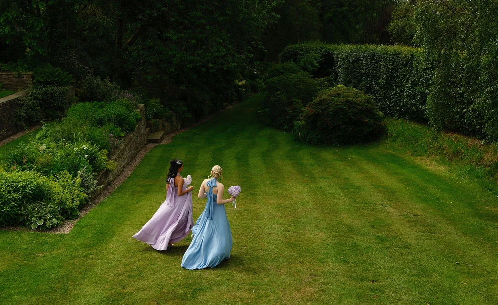 Two bridesmaids in pastel pink and blue dresses walk across a sprawling manicured lawn holding bouquets.