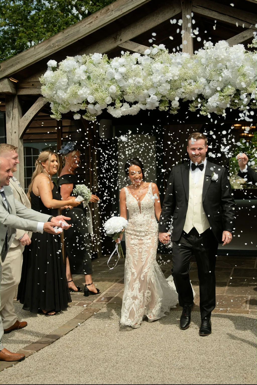 A joyful newlywed couple walks hand in hand through a shower of confetti beneath a grand white floral arch at a rustic barn venue.