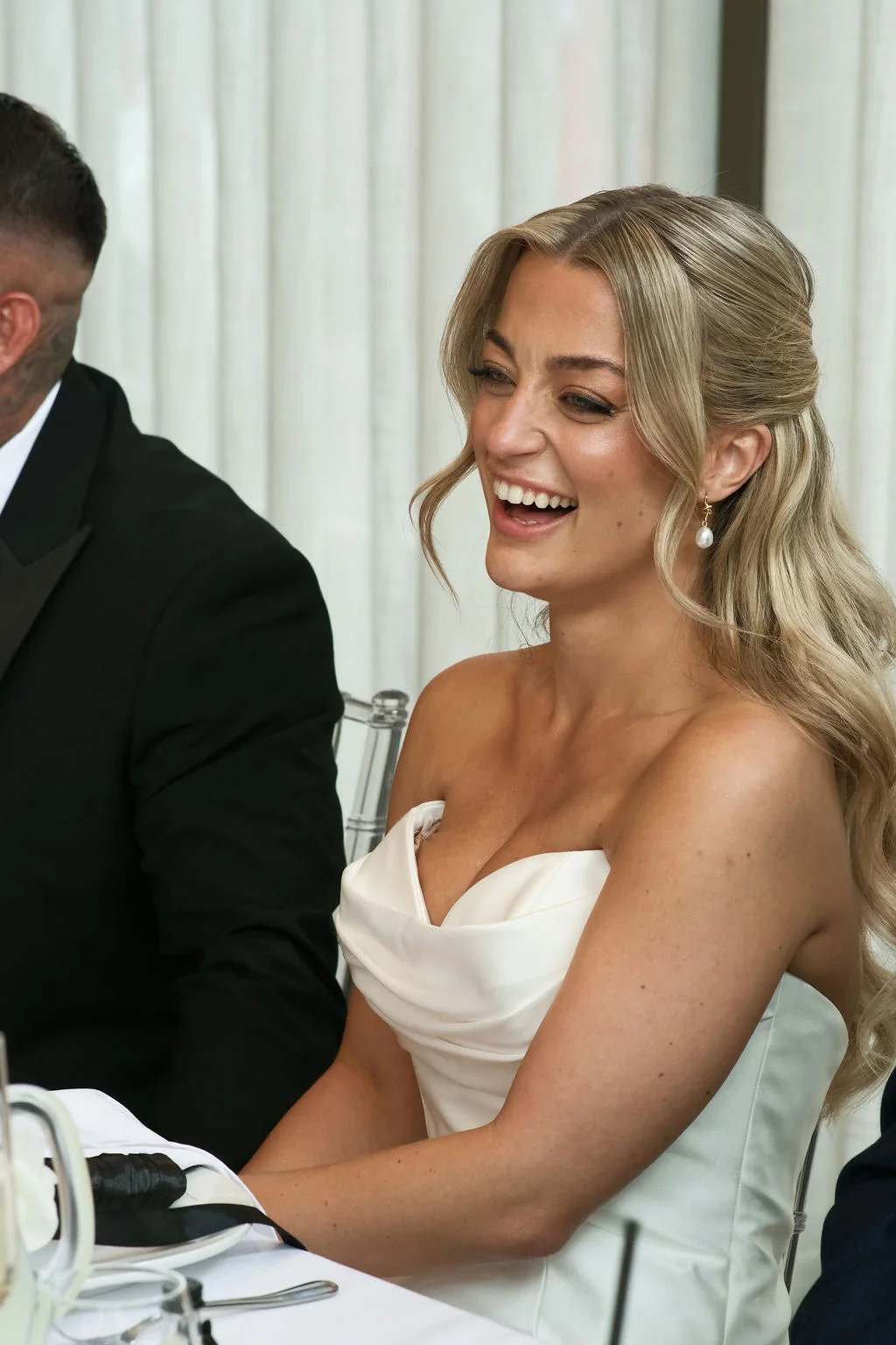 A smiling bride wearing a strapless draped gown laughs heartily during the wedding speeches at the top table.