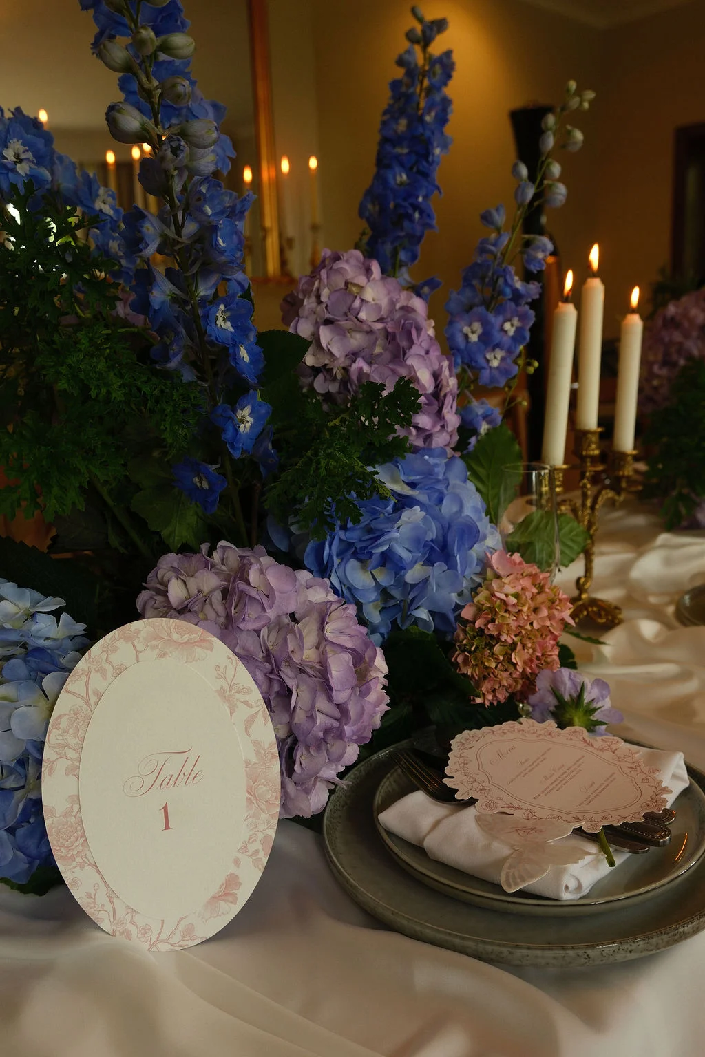 An elegant wedding tablescape features a magnificent floral centrepiece of blue delphiniums and purple and blue hydrangeas, accompanied by taper candles and a table number card for table one.