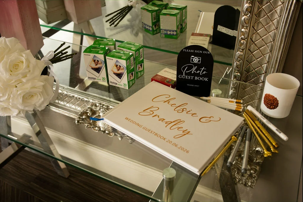 A close-up flat lay showcases a white wedding guest book with gold lettering, polaroid film boxes, pens, and a bouquet of white roses arranged on a glass table.