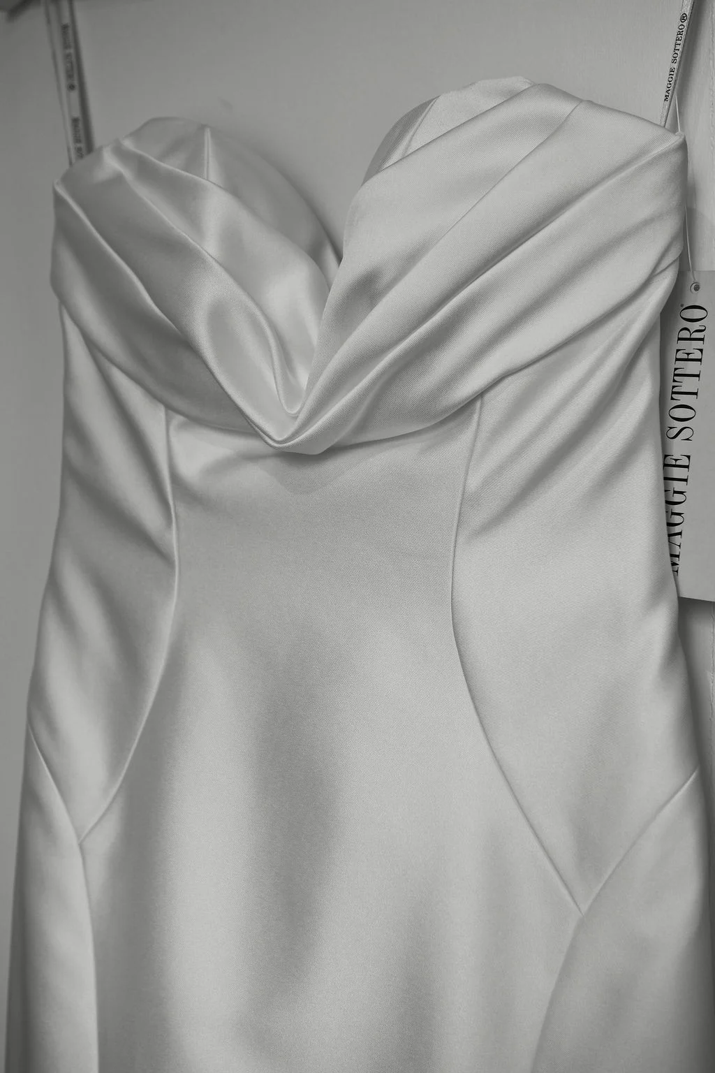 A close-up view of a structured satin wedding dress featuring a sweetheart neckline and ruched bodice, hanging indoors with a visible designer tag.