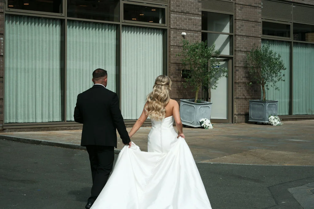 A newlywed couple walks hand in hand away from the camera along a city street, with the bride holding up her long train.