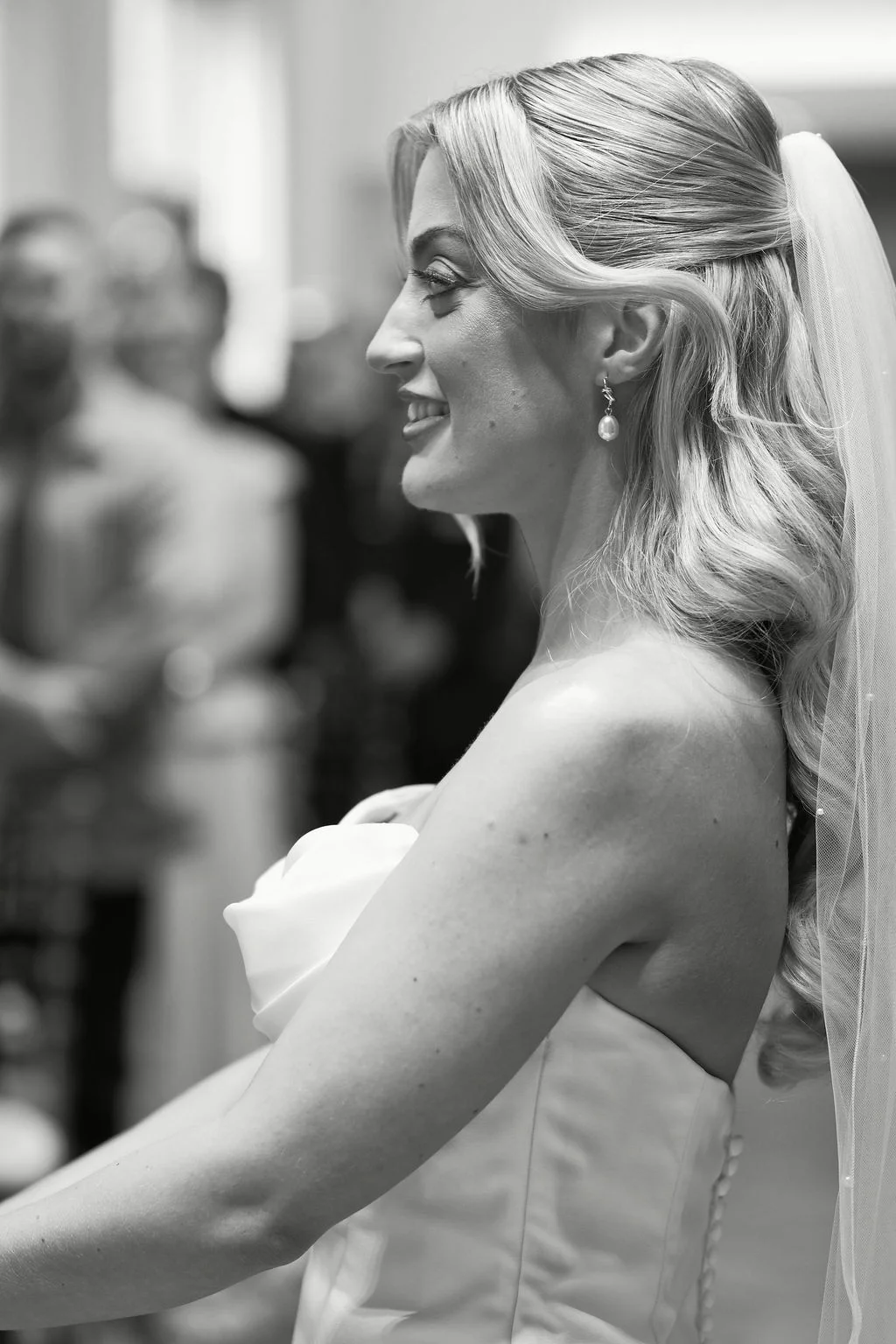 A beautiful bride smiling happily in profile view during her wedding ceremony, wearing a strapless gown with a half-up hairstyle and a delicate veil.