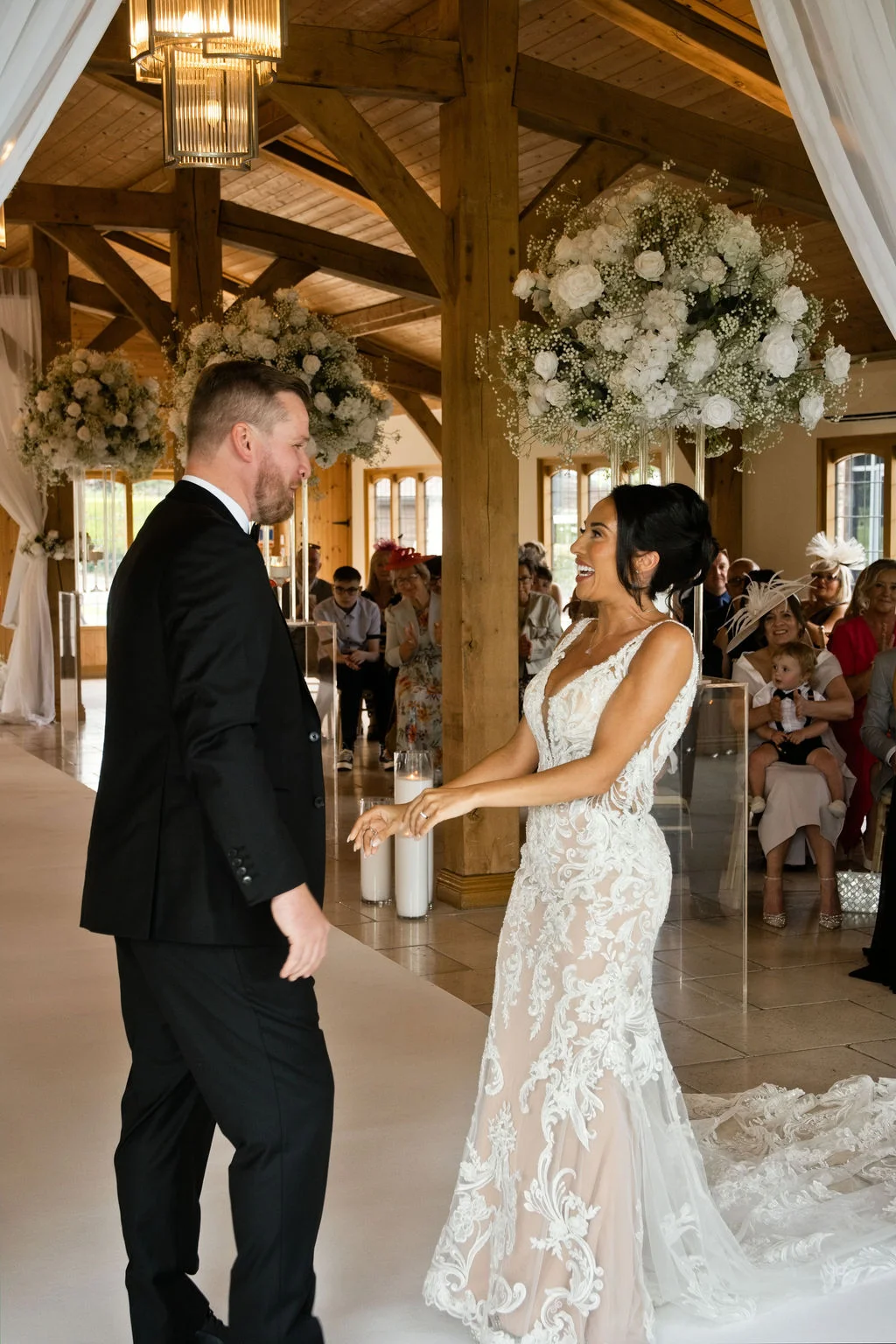 A newlywed couple share a joyful laugh at the end of the aisle inside a rustic oak-framed barn decorated with towering white floral arrangements.
