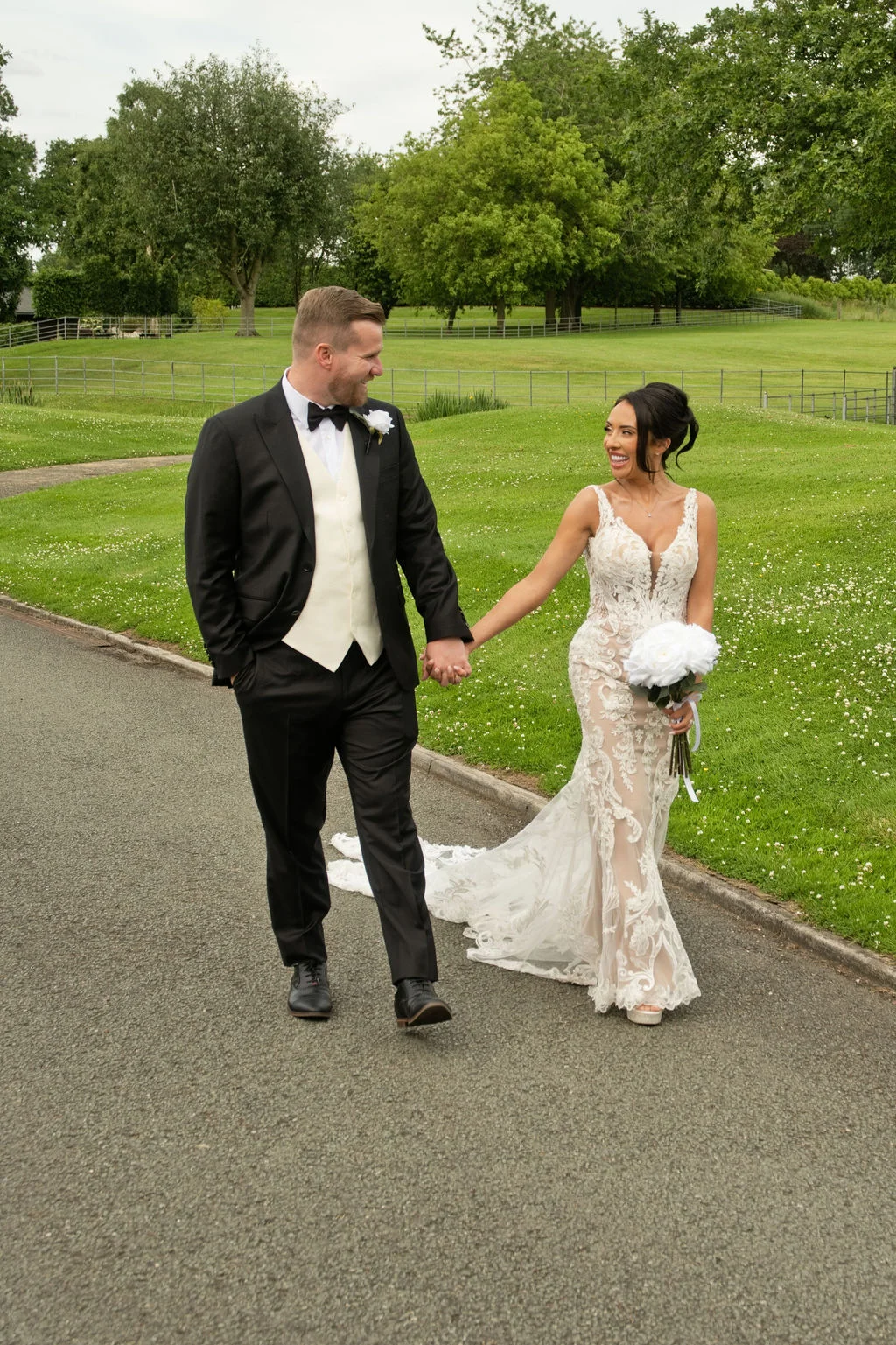 A newlywed couple walks hand in hand along a paved path bordered by green lawns, laughing and looking at each other outdoors.