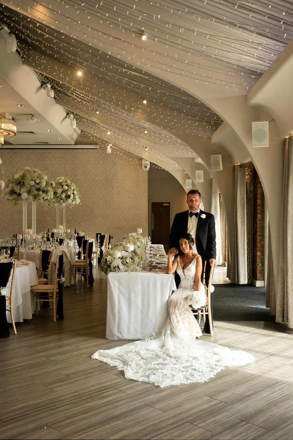 A newlywed couple poses in a grand hotel reception room featuring a vaulted ceiling draped with fairy lights, with the bride seated in a lace gown featuring a dramatic train beside the groom in a black tuxedo.