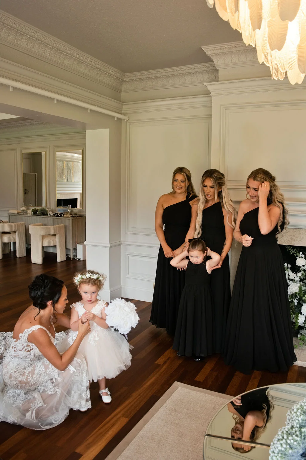 A bride in a detailed lace wedding dress kneels to help a young flower girl holding a white bouquet, while three bridesmaids in black dresses look on in an elegant manor house room.