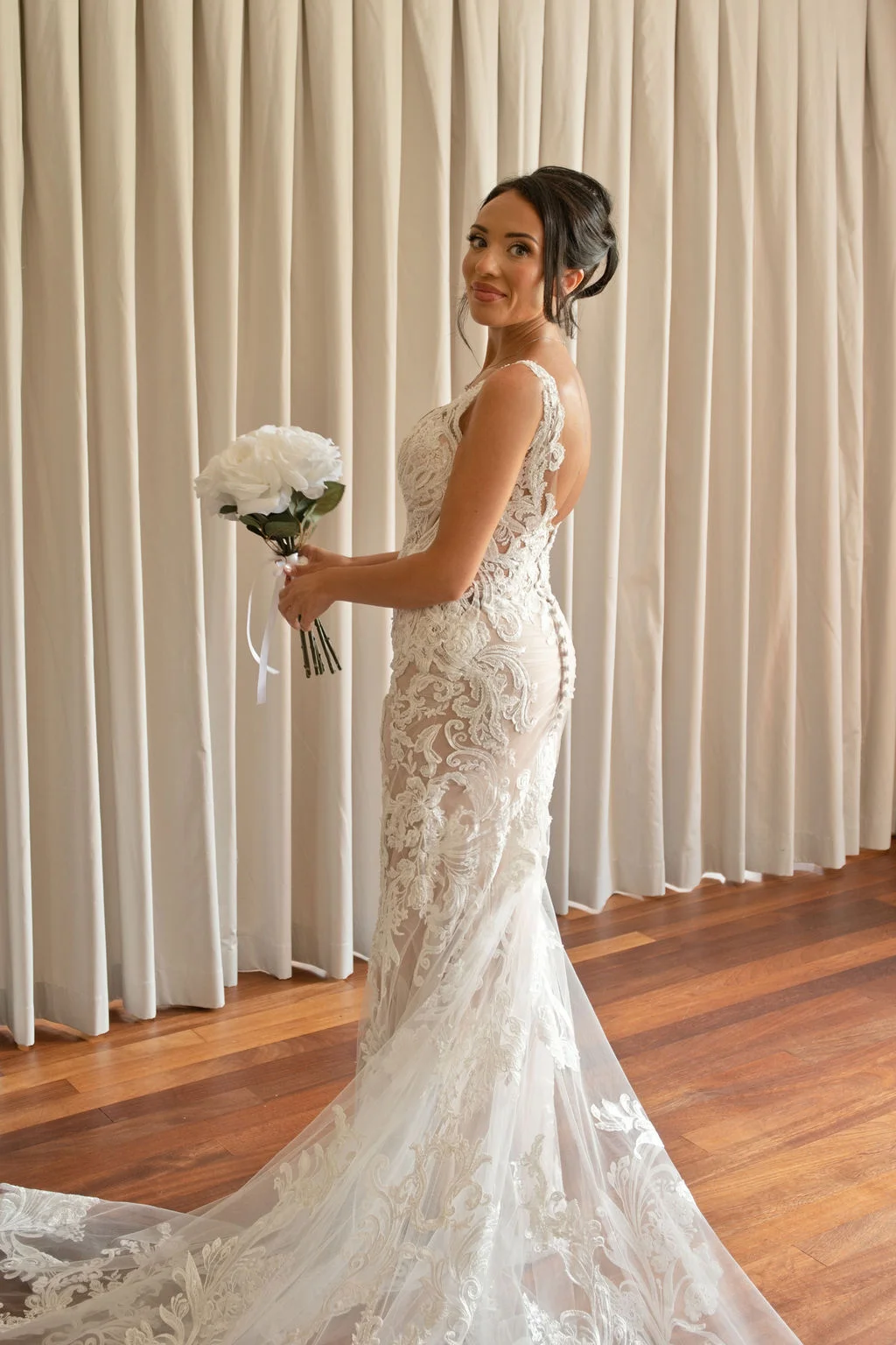 A stunning bride poses gracefully in a form-fitting lace wedding gown with an illusion back and train, holding a white rose bouquet against a neutral curtain backdrop.