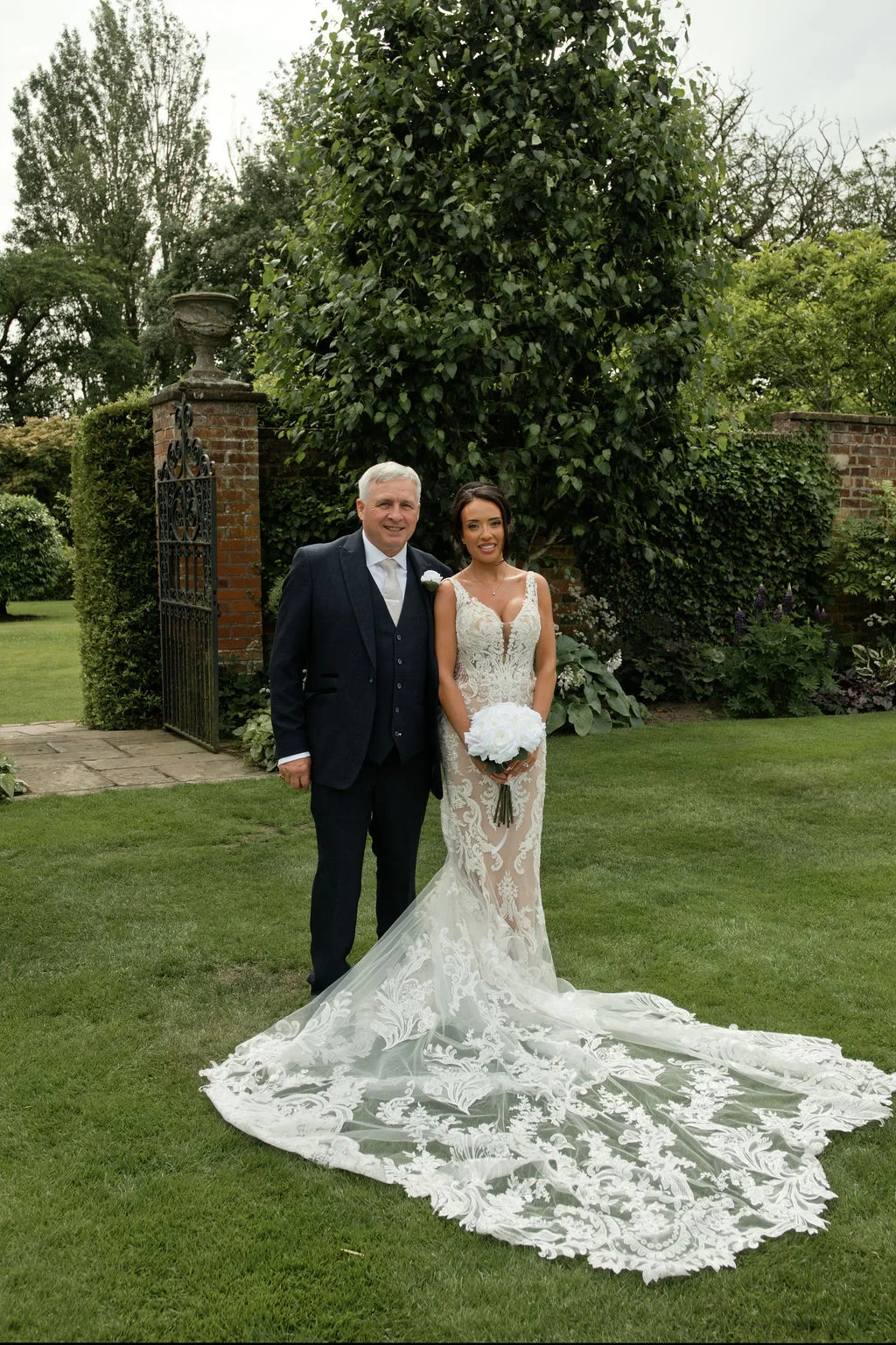 A bride in a lace gown with a dramatic train and her father pose together on a manicured lawn outside a walled garden.