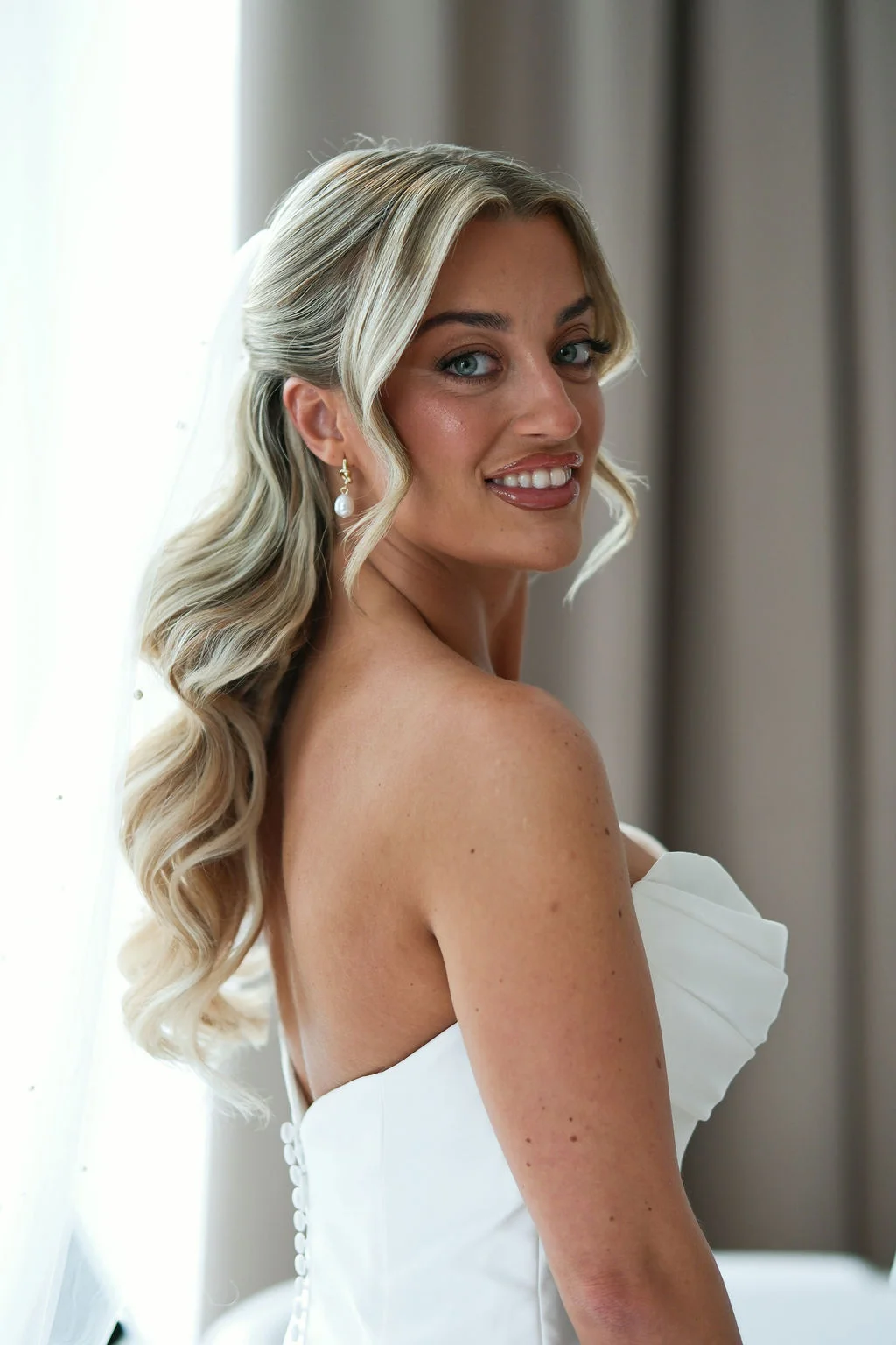 A stunning blonde bride with half-up waves and pearl drop earrings turns back to smile over her shoulder while wearing a strapless ruched wedding dress by a window.