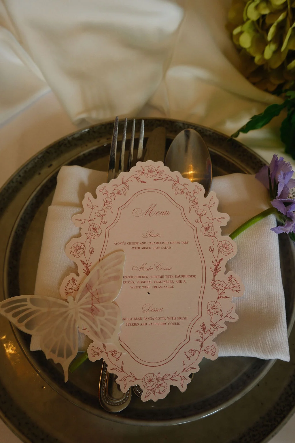 A detailed place setting features a decorative menu card adorned with delicate floral borders and a translucent butterfly resting on top, placed atop a folded white napkin and grey dinner plate.