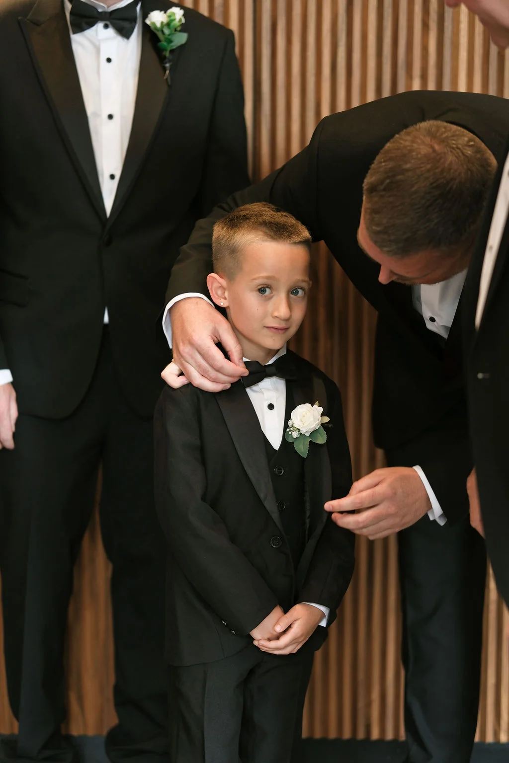 A young page boy dressed in a smart black tuxedo and bow tie is adjusted by a groomsman against a vertical wooden panelled backdrop.