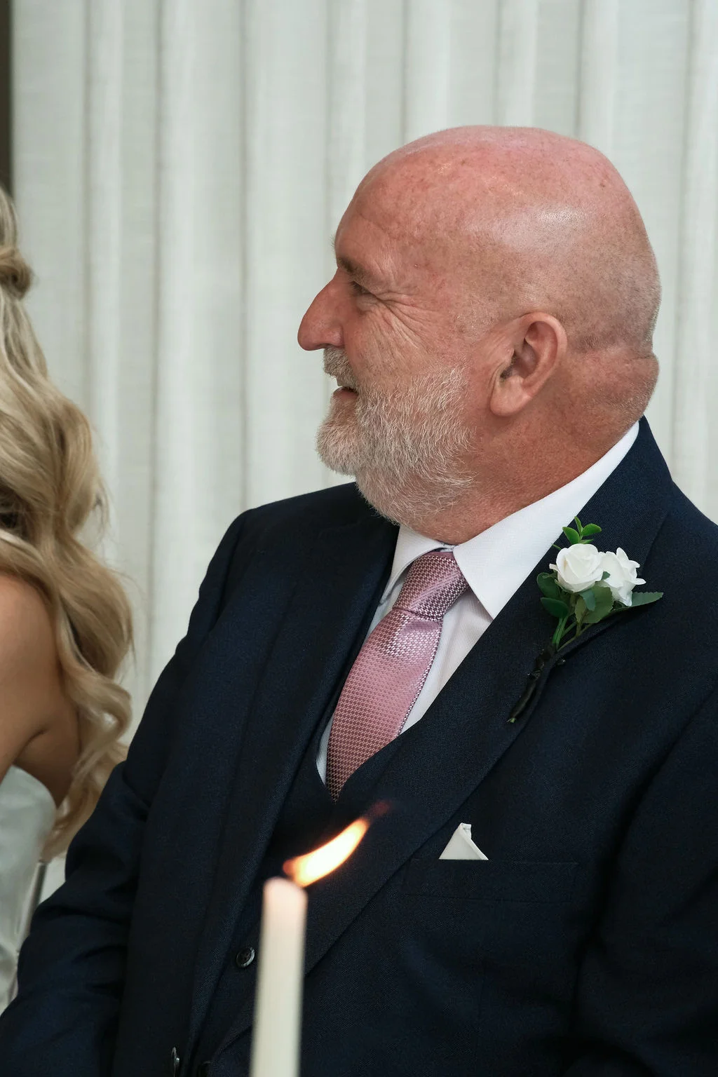 A smiling father of the bride wearing a navy three-piece suit, pink patterned tie, and white rose buttonhole looks on during wedding speeches.