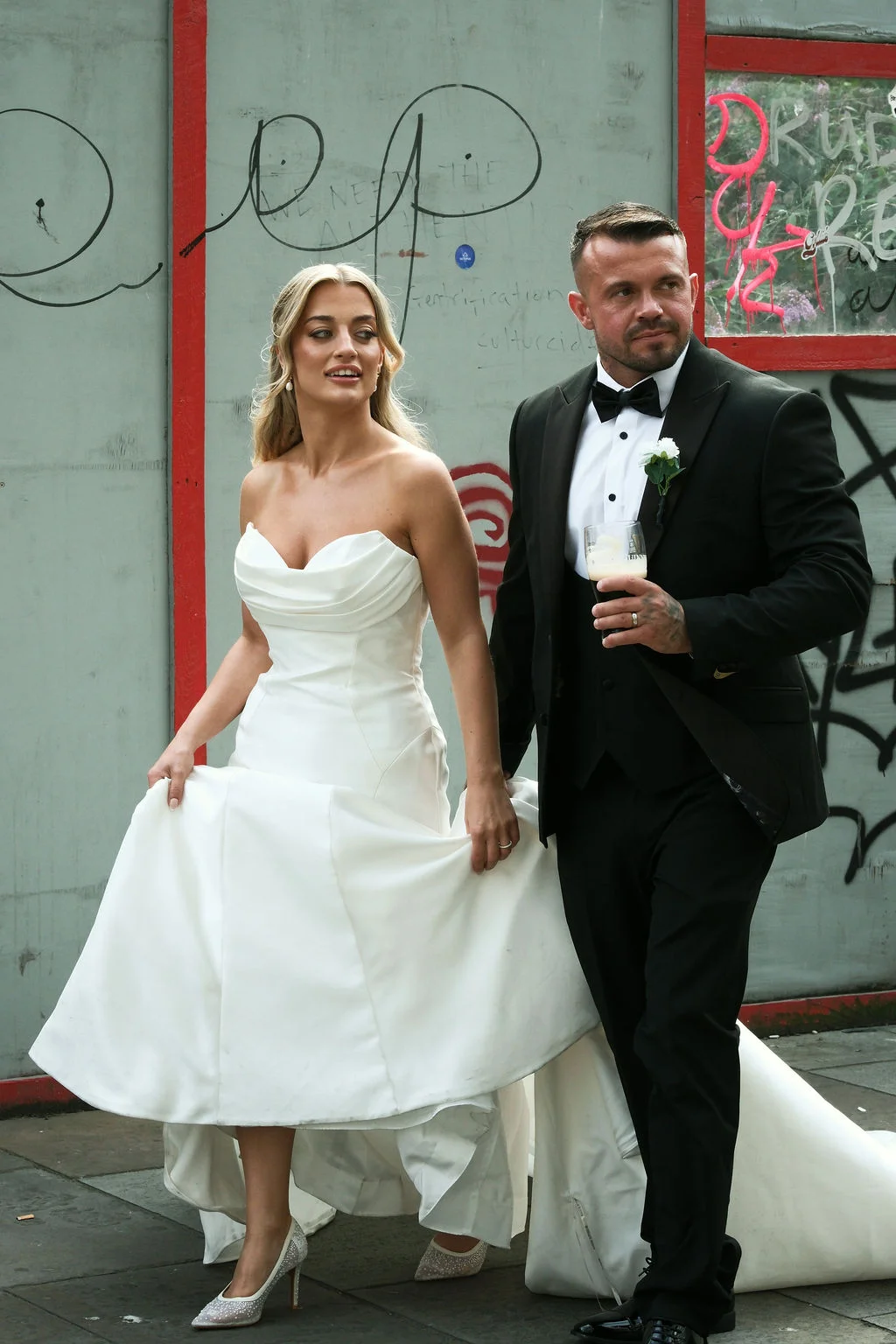 An urban bride and groom walk past a graffited wall, with the bride holding up her white satin a line gown while the groom holds a drink.