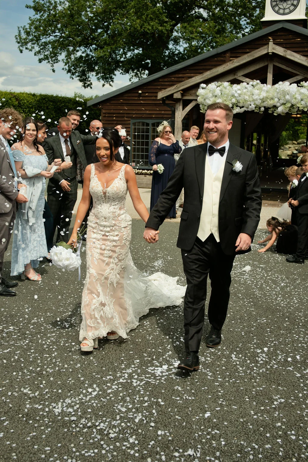 A joyful newlywed couple walks hand in hand down a gravel pathway as wedding guests shower them with white confetti outside a rustic barn venue.