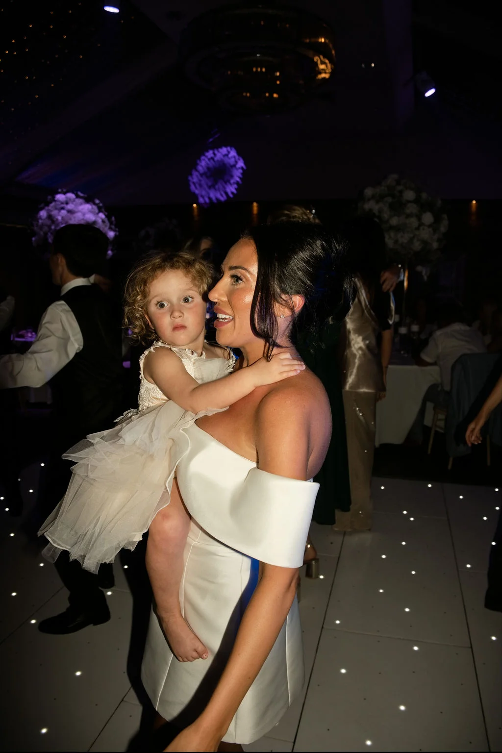 A guest holds a young flower girl in a ruffled dress on a glowing white dance floor during an evening wedding reception.