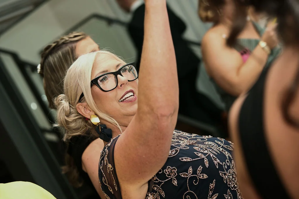 A guest at a wedding smiles joyfully with her arm raised in celebration while wearing a beaded dress and glasses.