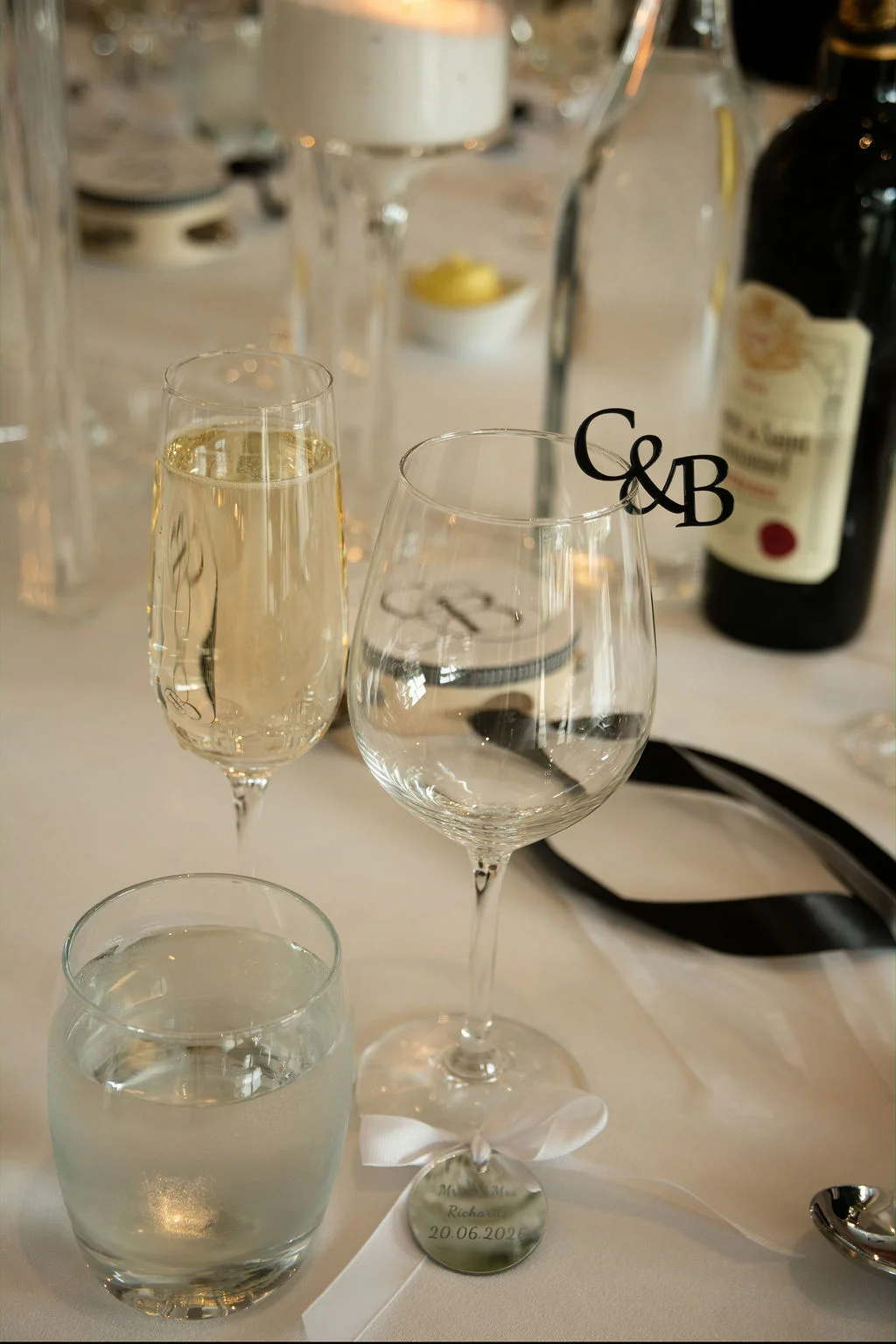 A close-up view of an elegant wedding tablescape featuring crystal glassware, a wine glass adorned with a custom monogram charm, and personalised favour tags resting on a white linen tablecloth.