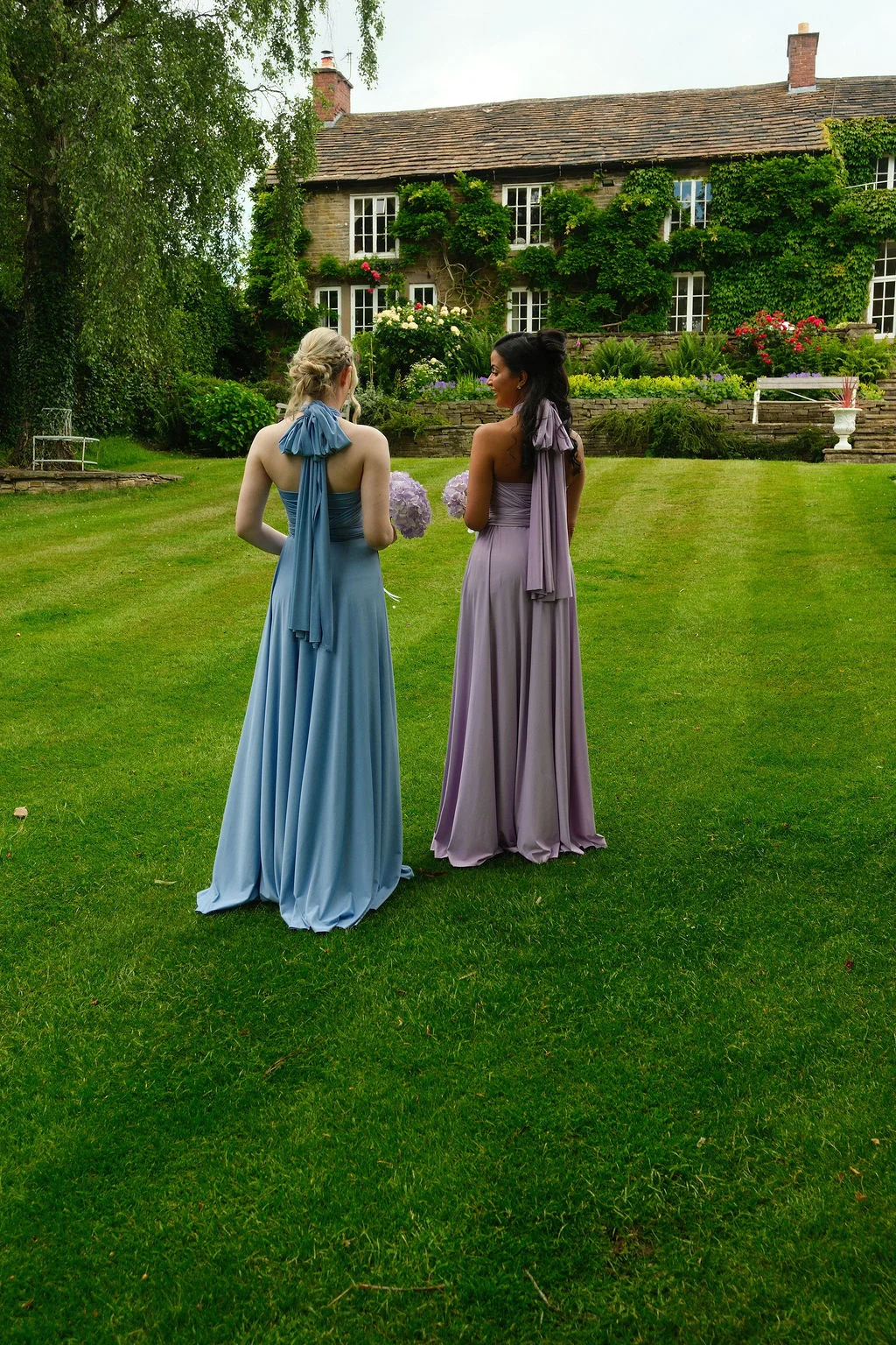 Two bridesmaids in pastel blue and pink halterneck dresses with bow-tied backs stand on a lush green lawn in front of an ivy-clad country house.