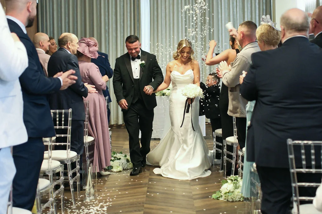 Newlyweds walk hand-in-hand down the aisle amidst a shower of white confetti while cheering guests applaud on either side.