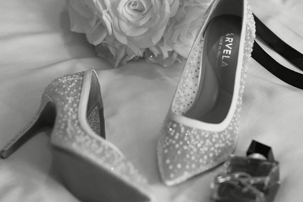 An elegant black and white flat lay features sparkling bridal heels arranged neatly alongside a white rose posy and a perfume bottle.