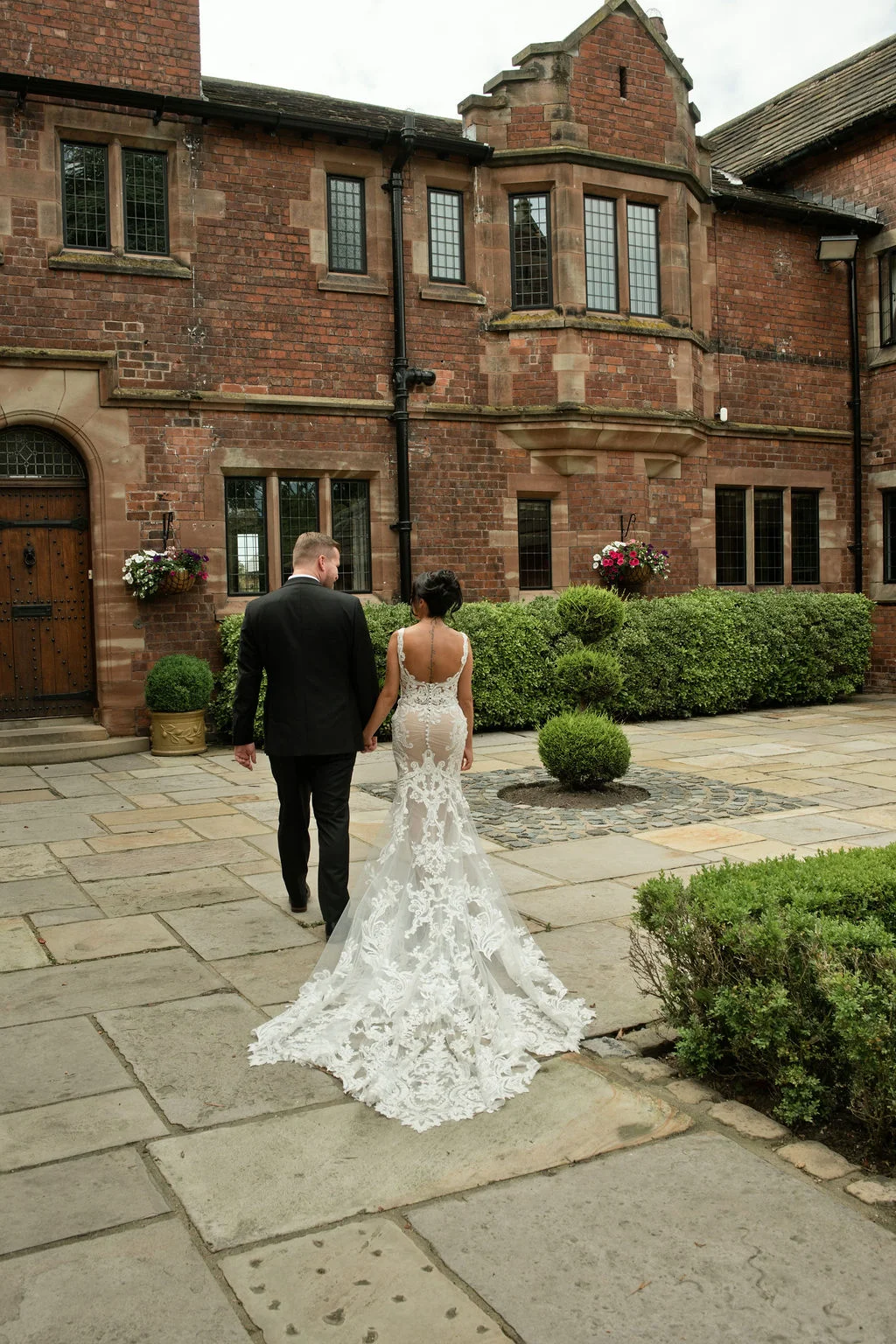 Newlyweds walk hand in hand away from the camera across a stone courtyard outside a historic red brick manor house.