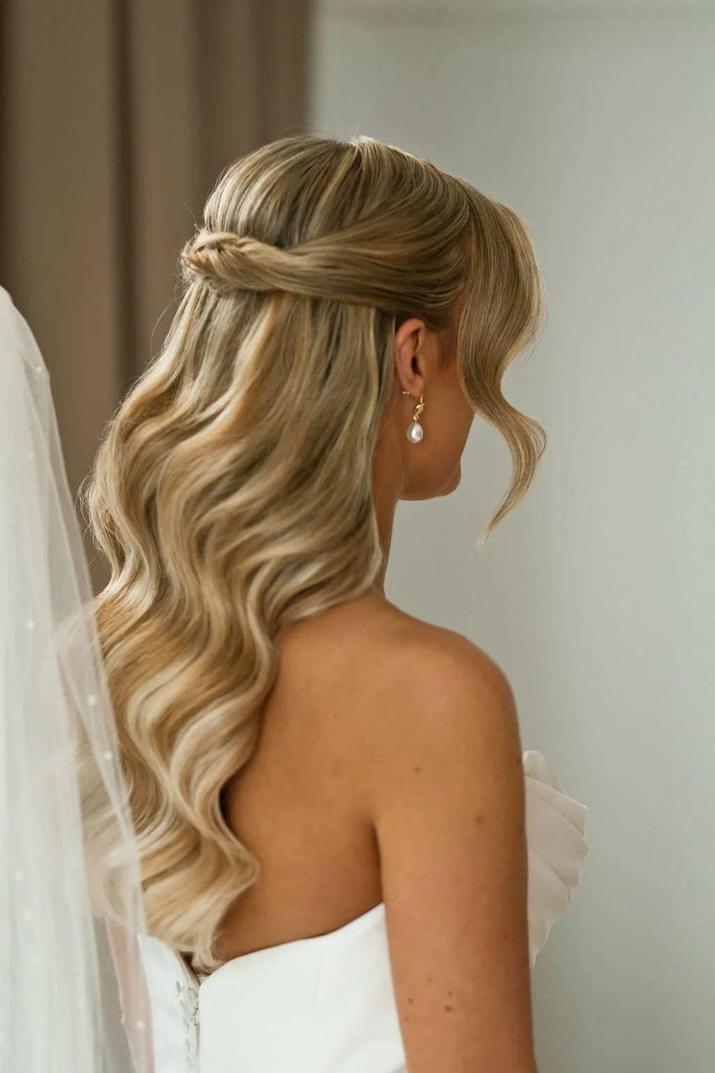 A close-up back view of a bride with long blonde hair styled in soft half-up waves, wearing pearl drop earrings and a strapless satin gown with a pearl veil.