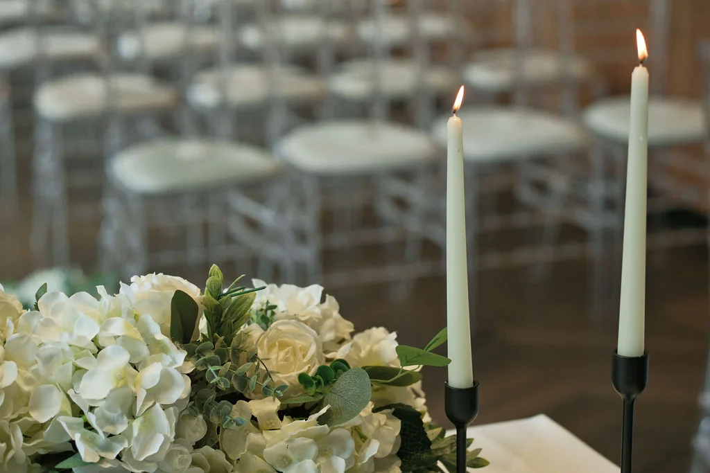 Lit white taper candles in black holders stand beside a lush arrangement of white roses and green foliage against a softly blurred background of ceremony seating.