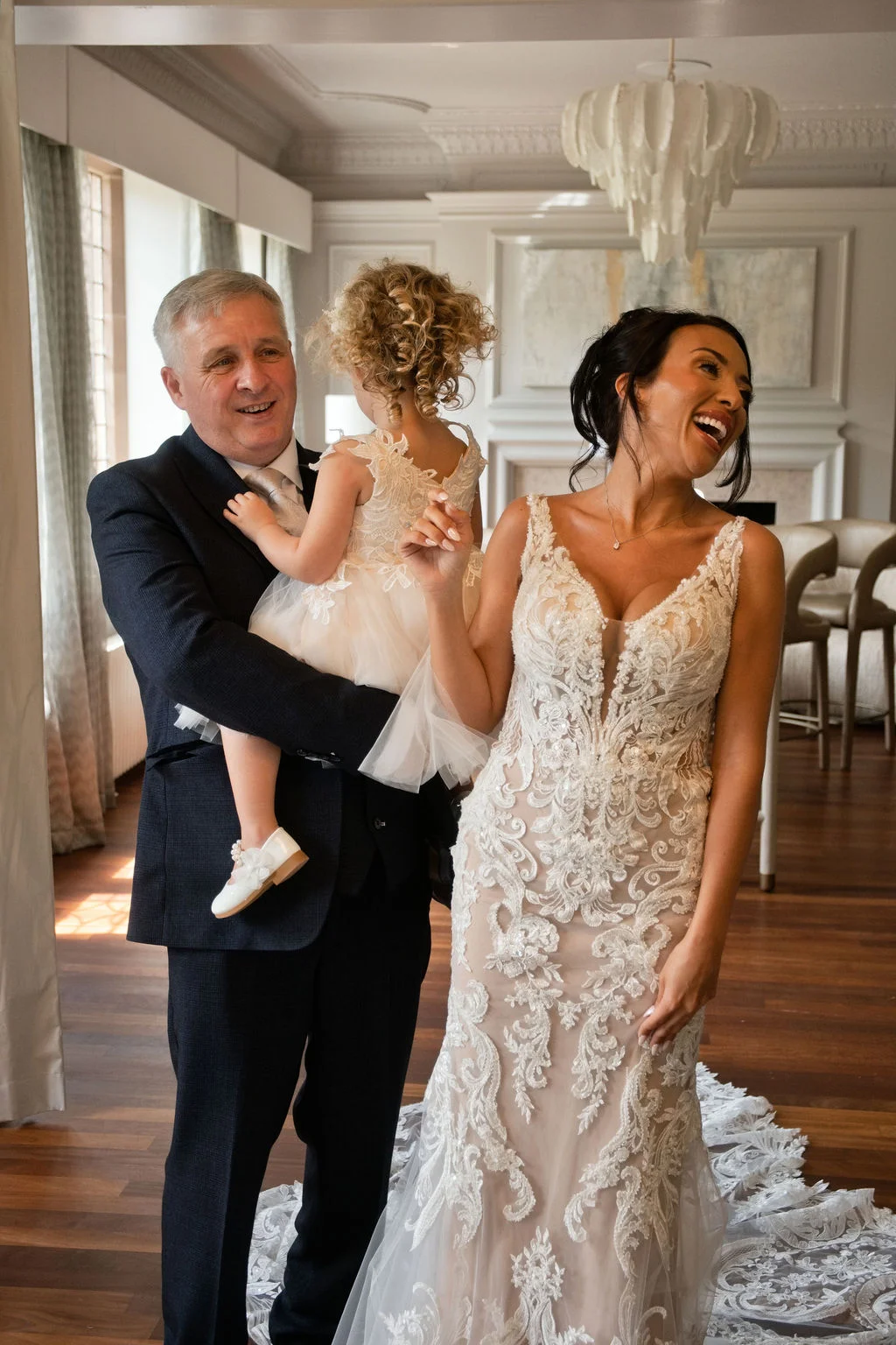 A joyful bride in a detailed lace gown shares a laughing moment with her father, who is holding a young flower girl in an elegant indoor venue.