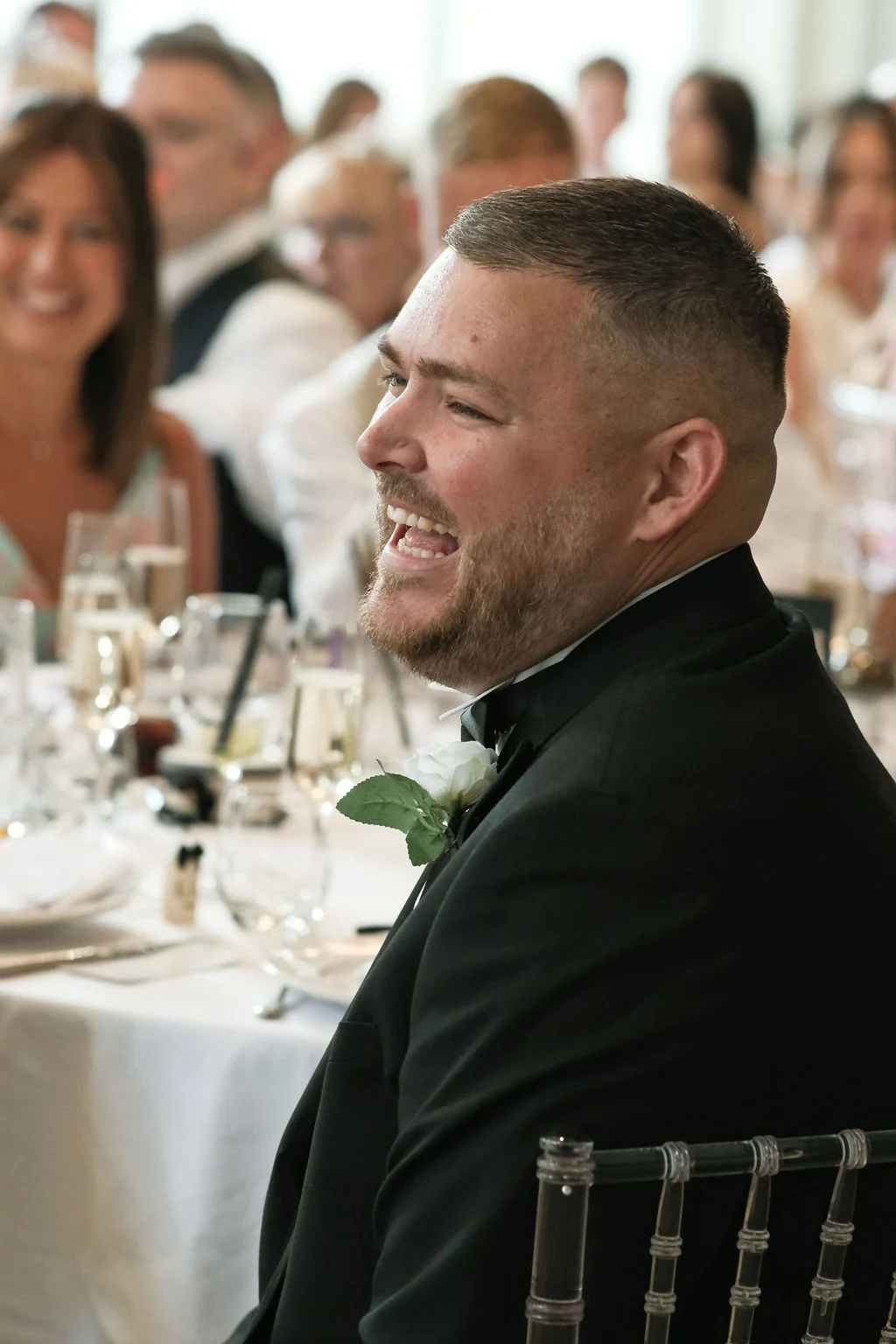 A smiling groom wearing a black tuxedo and white buttonhole laughs during the wedding speeches at a reception table.