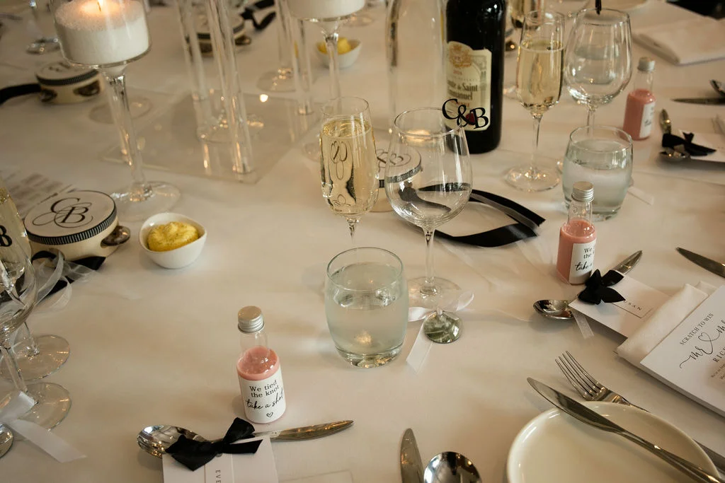 An elegant monochrome wedding tablescape featuring crisp white linen, personalised favour bottles, sleek black ribbon details, and glowing pillar candles.