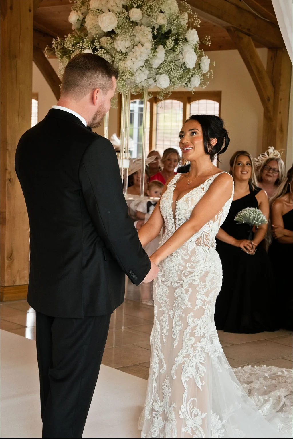 A newly married couple stand holding hands at the altar in an oak framed barn venue, smiling lovingly at each other during their wedding ceremony.