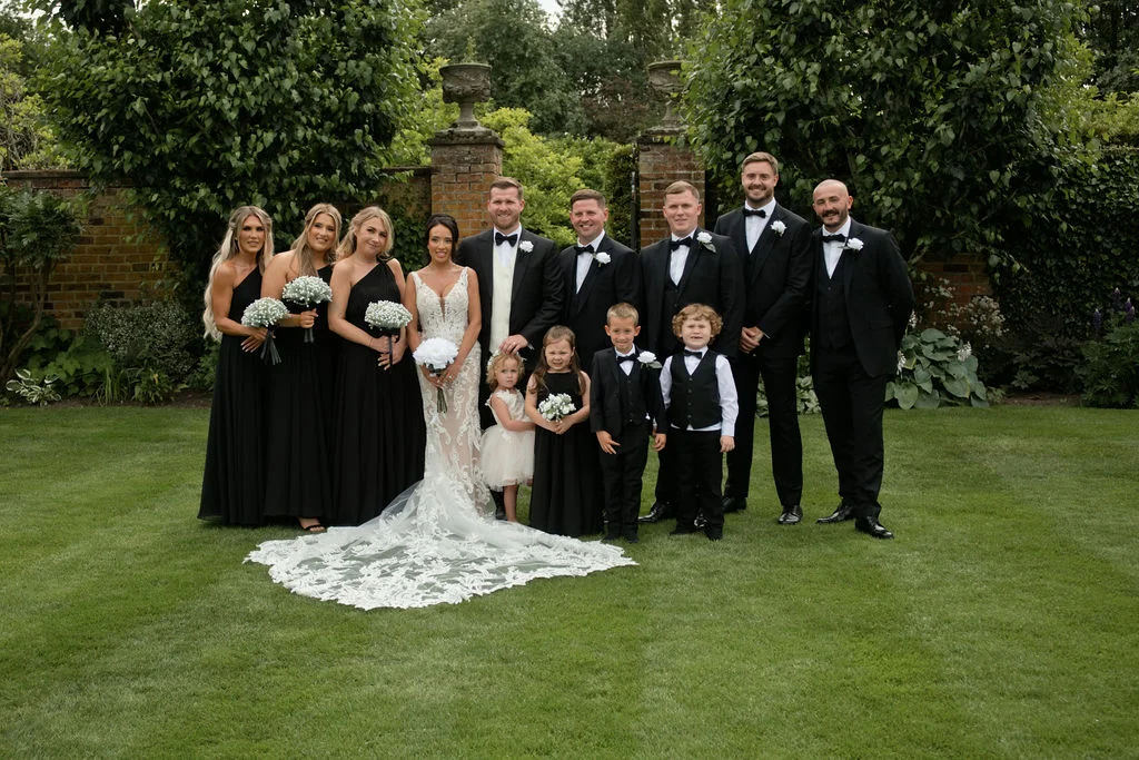 A bridal party dressed in elegant black attire poses on a manicured lawn outdoors, featuring bridesmaids in black gowns and groomsmen in black tuxedos.