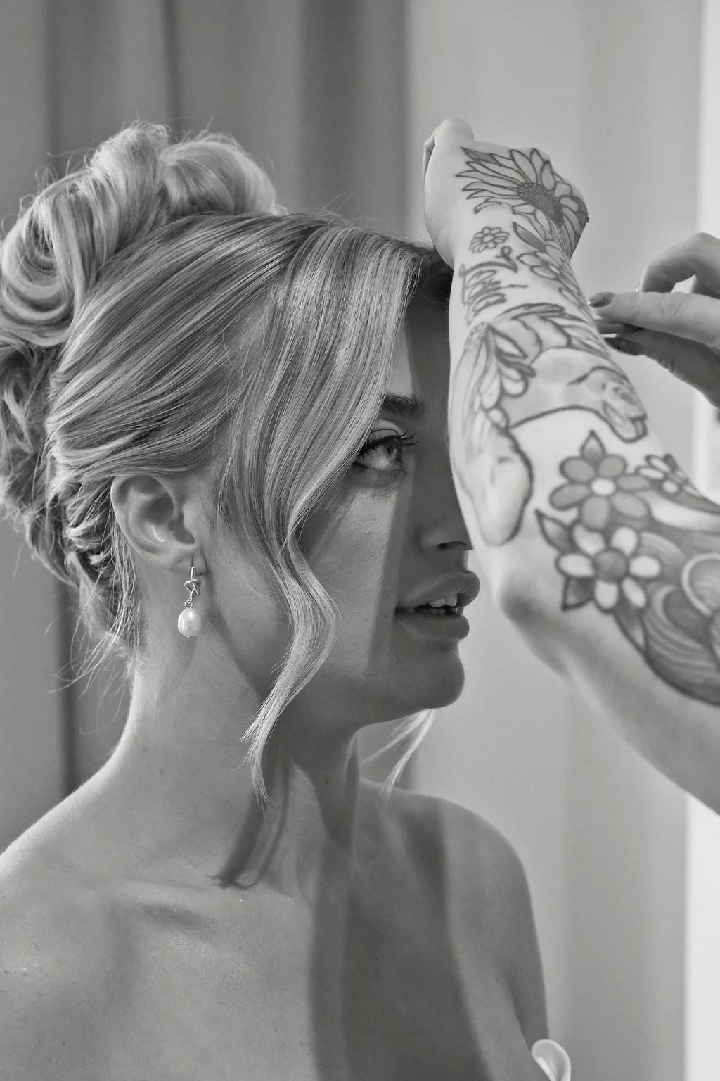 A close-up black and white shot captures a bride having her hair styled during morning preparations, wearing elegant pearl drop earrings and an updo.