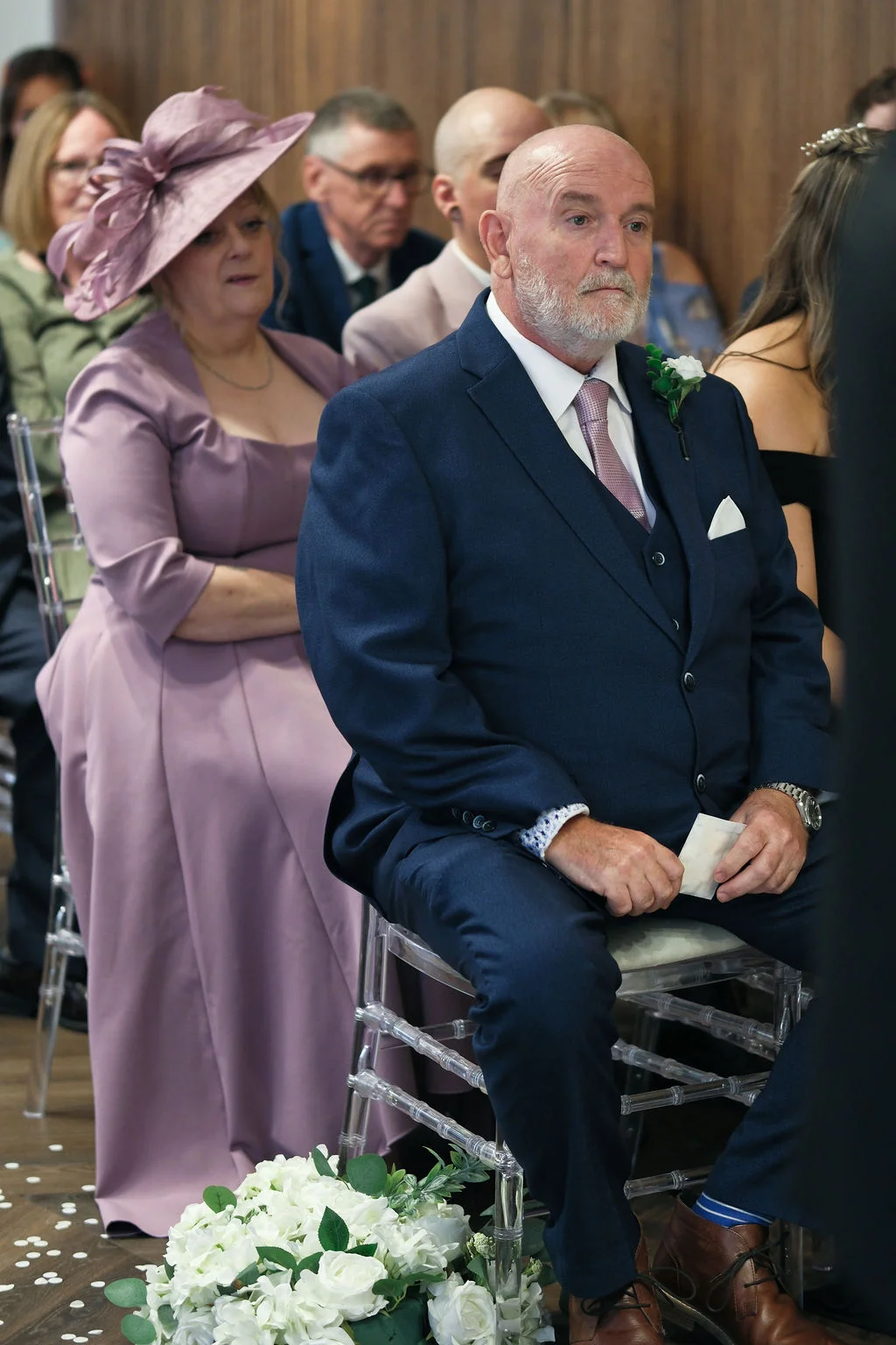 A mature gentleman in a tailored navy three-piece suit sits attentively on a clear acrylic chair among wedding guests during a ceremony.